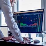 Man at desk looking at computer screen showing stock market trading data.