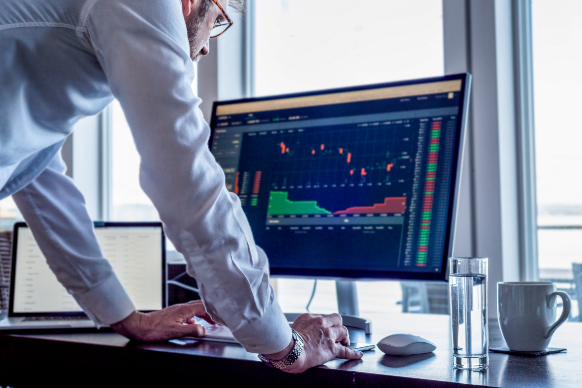 Man at desk looking at computer screen showing stock market trading data.